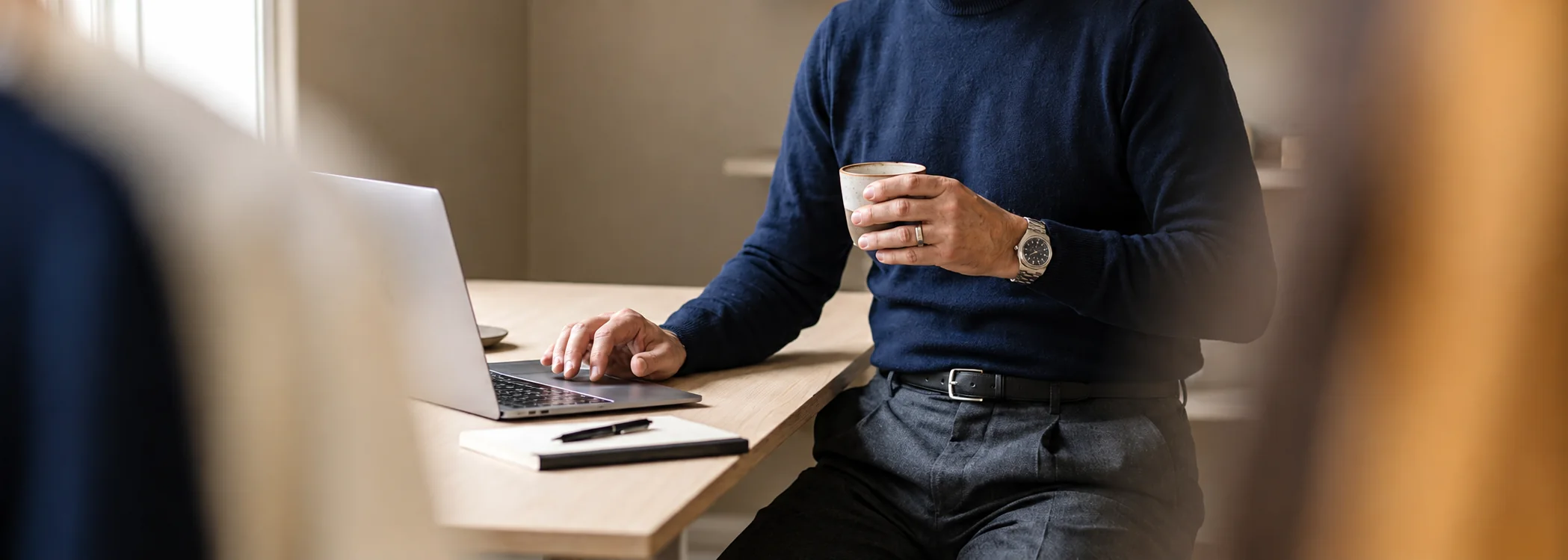 A person holding a cup while working on a laptop at a wooden table beside a window