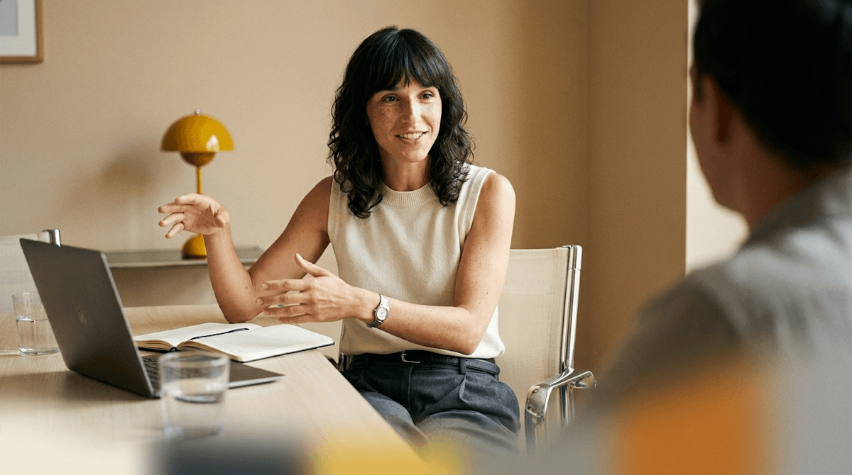 Woman discussing her work across a desk with a laptop and notebook