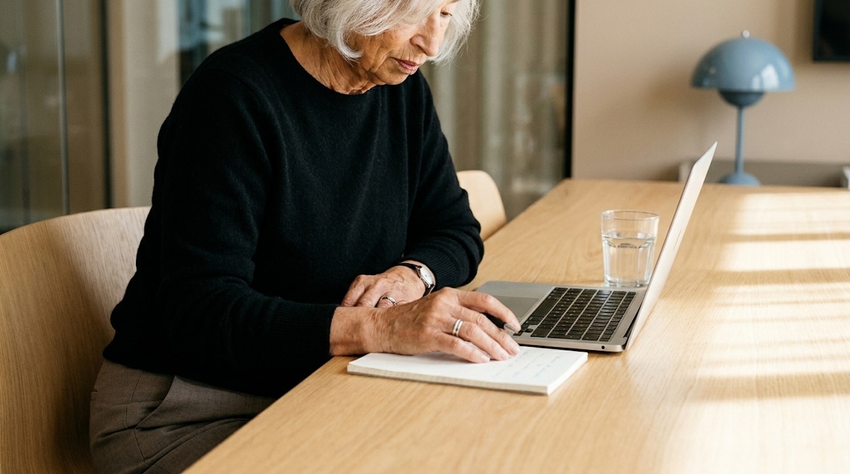 A woman with silver hair working with a notebook and laptop at a wooden table