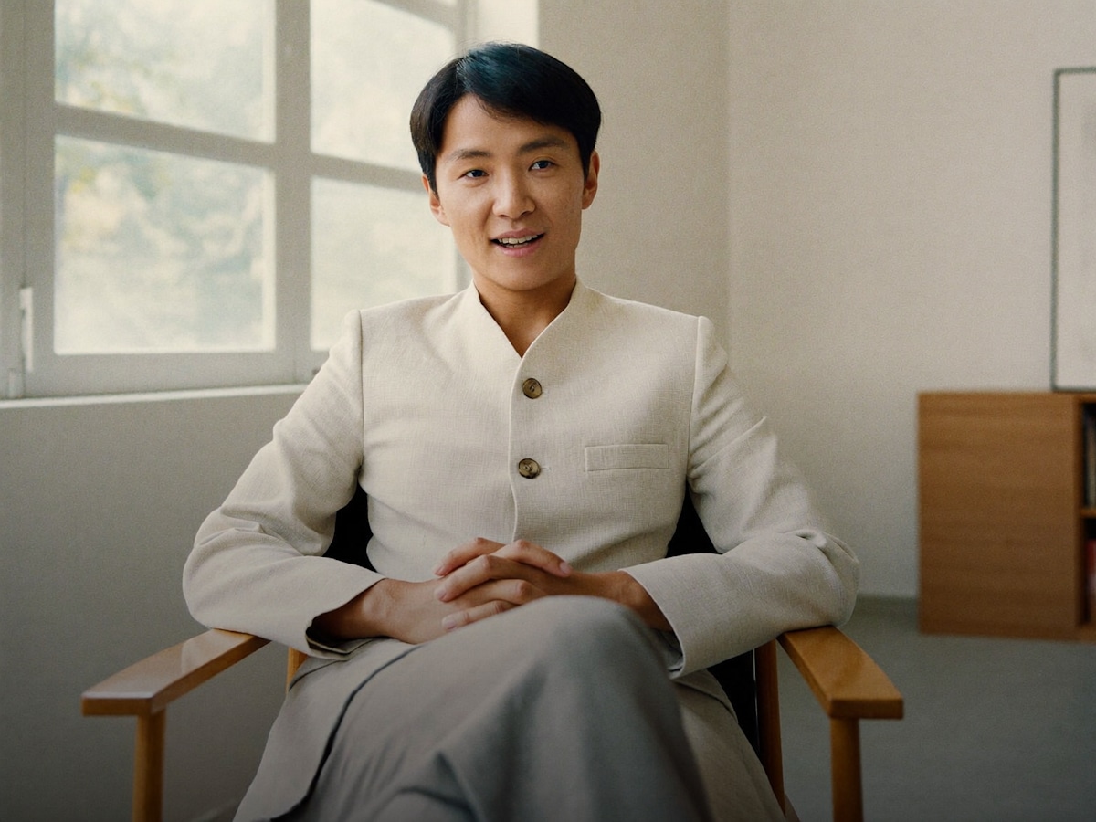 A man in a light jacket sitting in a wooden chair in a sunlit room
