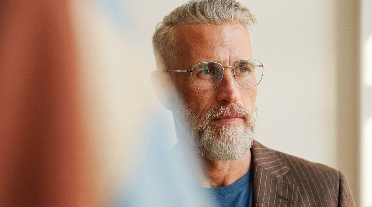 A man with glasses and a grey beard in a striped jacket beside a window