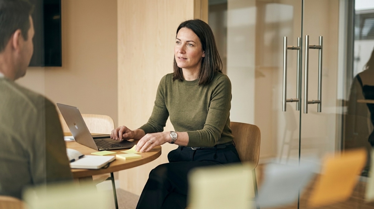 A woman in a green sweater discussing work across a table with an open laptop