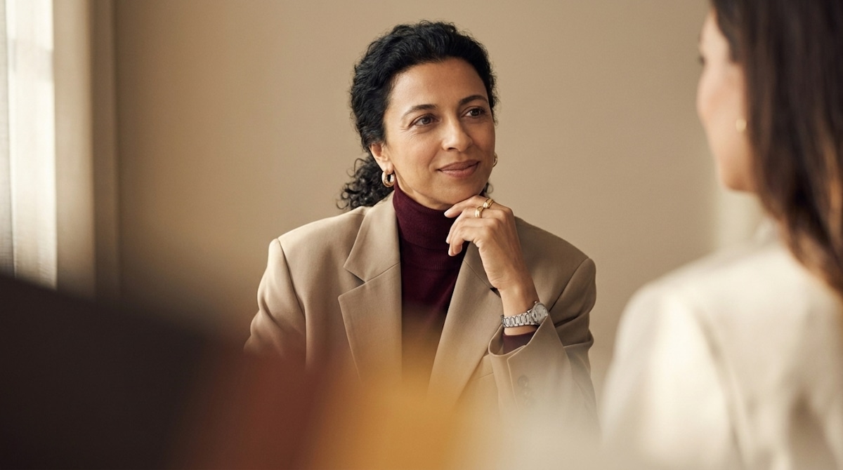 A woman in a beige blazer and burgundy sweater listening to someone across a table