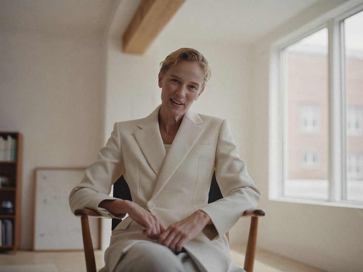 A woman in a cream suit seated in a wooden chair by large windows