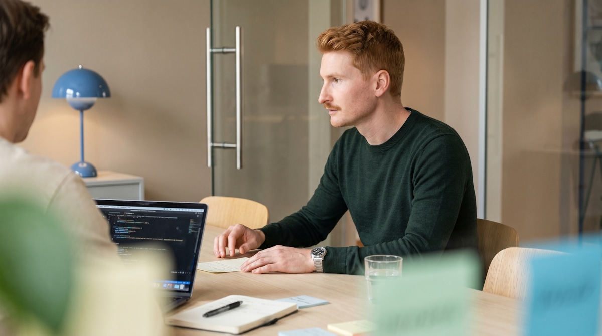 A man in a dark green sweater working at a table opposite a colleague with an open laptop