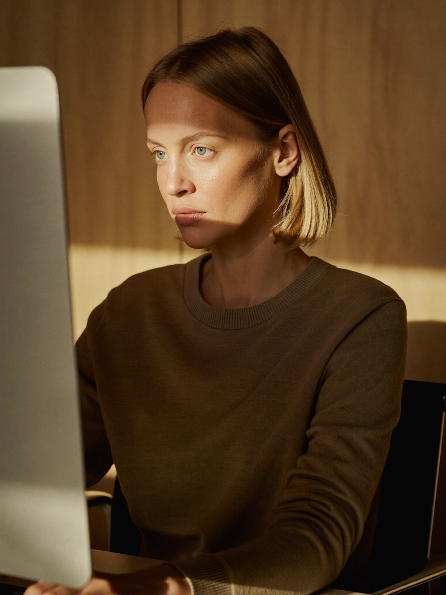 A woman in a brown sweater looking at a desktop monitor beside a sunlit wooden wall