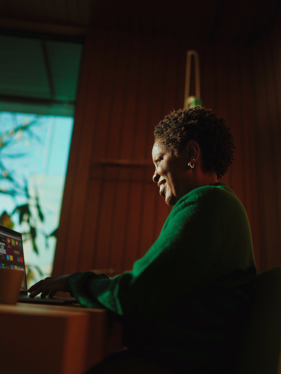 A woman in a green sweater working on a laptop beside a window