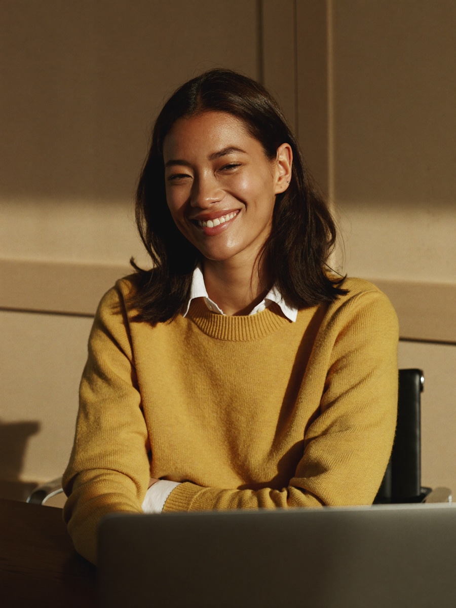 A smiling woman in a mustard sweater seated behind a laptop in warm sunlight