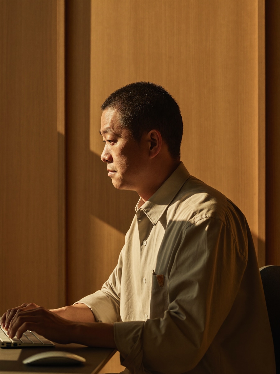 A man in a beige shirt typing at a wooden desk in afternoon sunlight