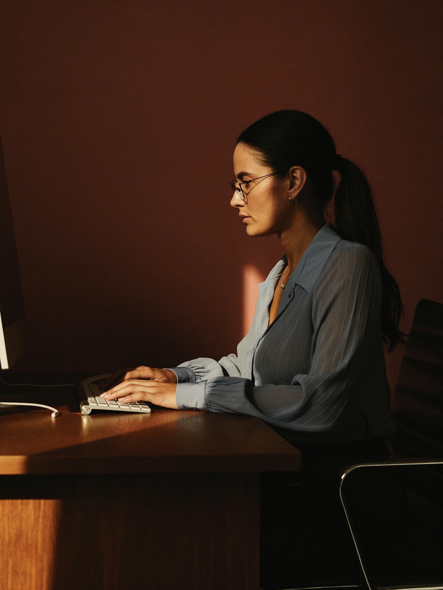 A woman with glasses and a blue blouse typing at a wooden desk